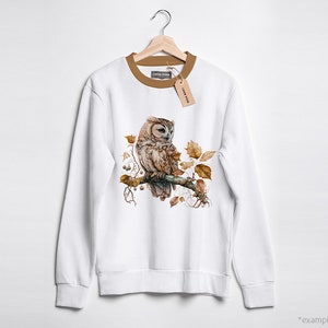 May include: White sweatshirt with a brown owl perched on a branch with autumn leaves. The owl has brown and white feathers and yellow eyes.