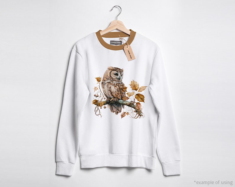 May include: White sweatshirt with a brown owl perched on a branch with autumn leaves. The owl has brown and white feathers and yellow eyes.