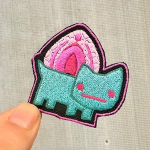 Può includere: Toppa ricamata raffigurante un gatto turchese stilizzato con dettagli rosa e un disegno rosa e viola sopra. La toppa ha un bordo nero e si trova su uno sfondo grigio chiaro.