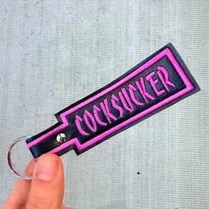May include: Black leather keychain with the word "COCKSUCKER" embroidered in pink. The keychain has a pink border and a silver key ring.