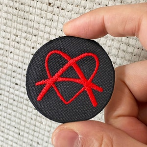 Op de afbeelding: Een zwarte stoffen patch met een rood geborduurd anarchiesymbool in een hart.