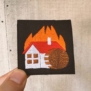 Op de afbeelding: Geborduurde patch met een huis in brand. Het huis heeft een witte behuizing, een rood dak en bruine ramen. Oranje en gele vlammen omringen het huis. Een bruin wiel staat naast het huis, alles op een donkerbruin vierkant.