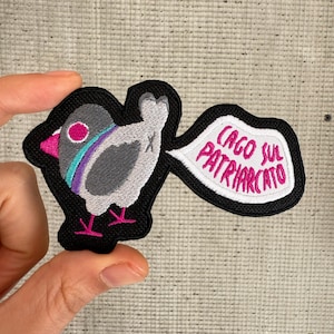 Peut inclure: Patch brodée thermocollante représentant un oiseau de dessin animé avec une bulle de dialogue qui dit "CAGO SUL PATRIARCATO". L'oiseau est gris avec des accents roses, violets et bleus. La bulle de dialogue est blanche avec du texte rose.