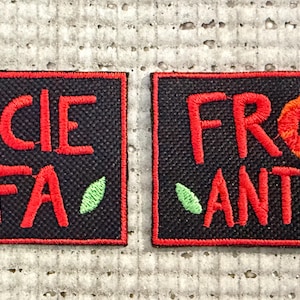 Op de afbeelding: Twee zwarte rechthoekige patches met rood geborduurde tekst en bloemmotieven. Eén patch leest "SOCIE IFA" en de andere "FRO ANTI". Beide patches hebben rode randen, rode tekst en groene blad- en steeld details.