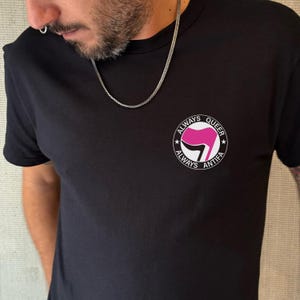 May include: Black t-shirt with a circular graphic on the chest. The graphic features a pink and white design with the words "ALWAYS QUEER ANTIFA" in a circular arrangement. The shirt has short sleeves and a crew neck.