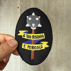 Può includere: Toppa ovale nera con una mazza grigia con accenti bianchi su un manico marrone. La toppa include banner gialli e blu con il testo italiano "A TUO RISCHIO E PERICOLO".