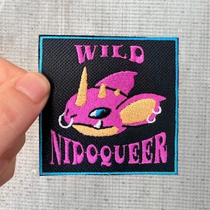 May include: A black embroidered patch with a pink cartoon creature with yellow ears and a blue eye. The text "WILD NIDQUEER" is embroidered in pink on the patch.