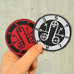 Peut inclure: Deux écussons brodés circulaires. L'un est rouge avec des accents noirs, l'autre est noir avec des accents blancs. Les deux présentent un symbole central et du texte environnant. Les écussons sont tenus par une main.