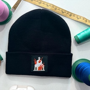 Op de afbeelding: Zwarte beanie met een vierkante patch met een kerk in brand, met rode en witte accenten. De beanie is omgeven door klosjes garen en een meetlint, wat wijst op een ambachtelijk of naai-thema.