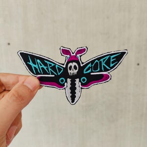May include: Black and white embroidered patch of a moth with a skull in the center. The moth's wings have the word "HARDCORE" written in turquoise and pink.