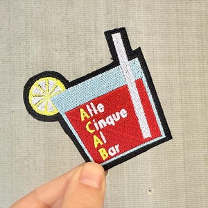 Può includere: Toppa ricamata raffigurante un cocktail con una fetta di limone e una cannuccia. La toppa riporta le parole "Alle Cinque Al Bar" in bianco e giallo su sfondo rosso, con una parte superiore blu e un bordo nero.