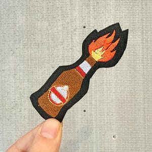 Può includere: Toppa ricamata con un disegno di cocktail Molotov. La toppa raffigura una bottiglia marrone con un emblema rosso e bianco, un collo bianco e fiamme arancioni e gialle. La toppa ha un bordo nero.
