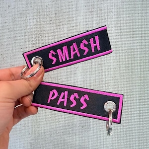 Può includere: Due portachiavi neri con testo ricamato rosa che dice "SMASH" e "PASS". I portachiavi hanno un bordo rosa e anelli argentati. Lo sfondo è una superficie strutturata grigio chiaro.