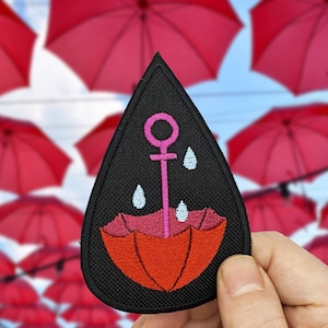 Può includere: Toppa ricamata nera con un'ancora rosa e un ombrello rosso con gocce di pioggia. L'ancora ha un cerchio in cima e l'ombrello ha un design a strisce rosa e bianco.