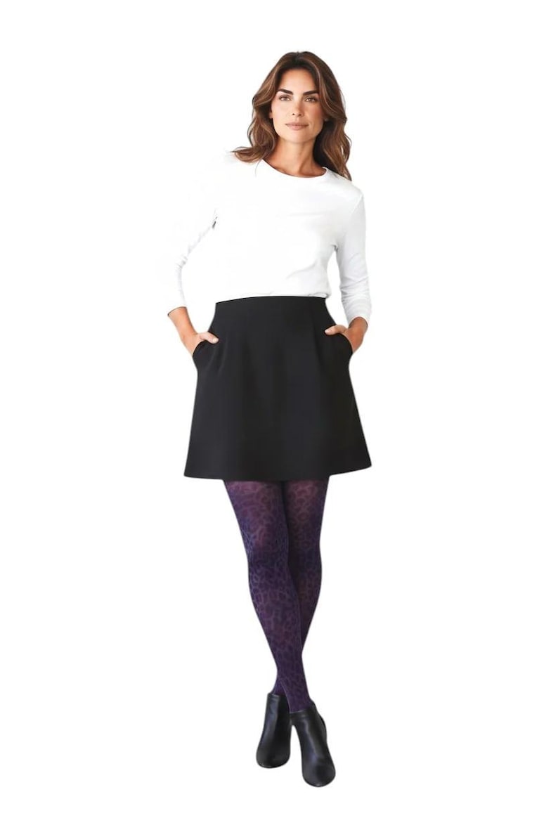 A woman standing with hands in pockets, wearing a white long-sleeved top, black mini skirt, black ankle boots, and deep purple leopard print patterned tights.
