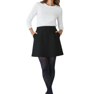 A woman standing with hands in pockets, wearing a white long-sleeved top, black mini skirt, black ankle boots, and dark navy blue leopard print patterned tights.