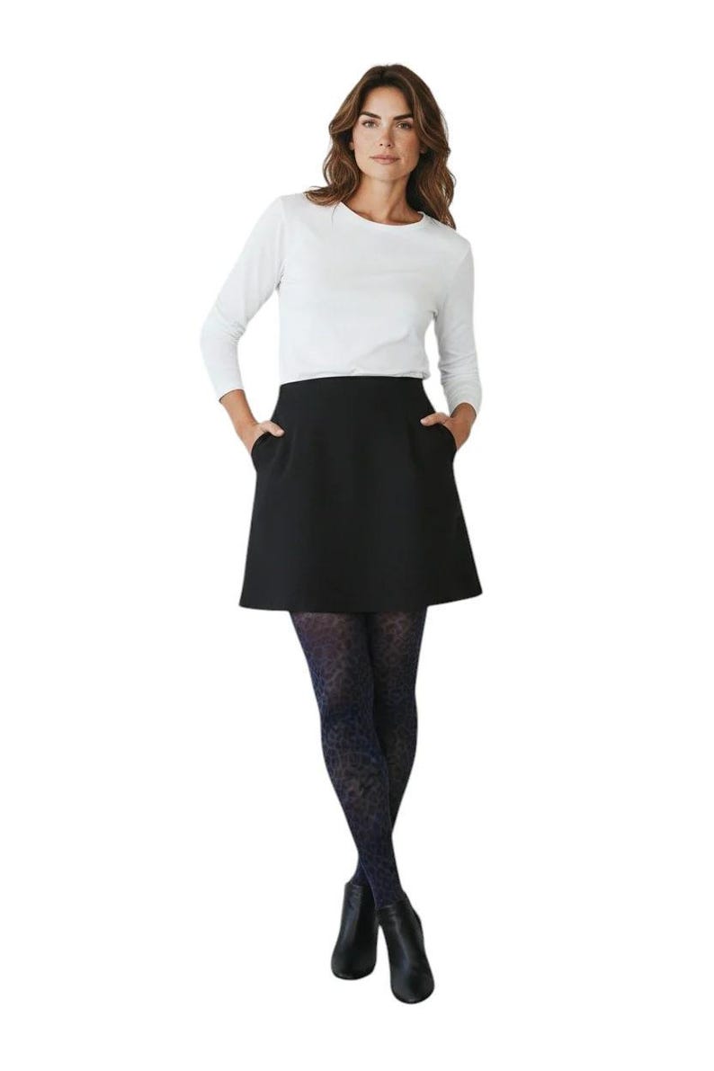 A woman standing with hands in pockets, wearing a white long-sleeved top, black mini skirt, black ankle boots, and dark navy blue leopard print patterned tights.