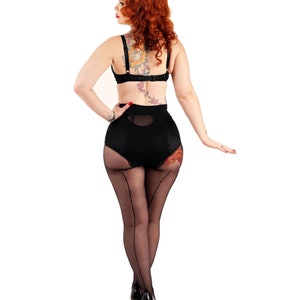 Op de afbeelding: Een vrouw die zwarte lingerie draagt met een bloementaattoo op haar rug en dij. Ze draagt zwarte kousen met een naad aan de achterkant van de benen en zwarte hakken.