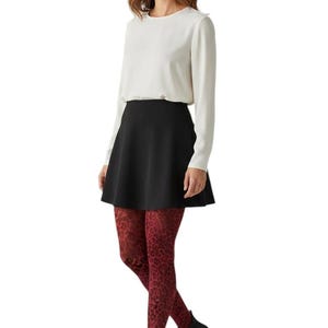 A woman standing, wearing a white long-sleeved top, black mini skirt, black ankle boots, and dark burgundy leopard print patterned tights.