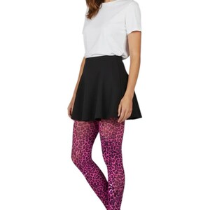 A woman standing with hands in pockets, wearing a white long-sleeved top, black mini skirt, black ankle boots, and dark neon pink leopard print patterned tights.
