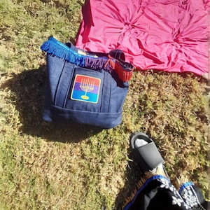 May include: A blue denim tote bag with a rainbow menorah patch and blue and purple fringe. The bag is sitting on green grass with a pink blanket in the background.