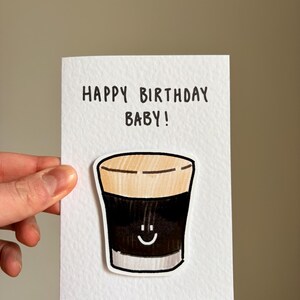 Puede incluir: Una tarjeta de felicitaci&oacute;n blanca con el texto "HAPPY BIRTHAY BABY!" en negro. La tarjeta presenta un dibujo de dibujos animados de una bebida oscura con una cara sonriente. La bebida tiene una parte superior de color canela y un cuerpo marr&oacute;n oscuro.