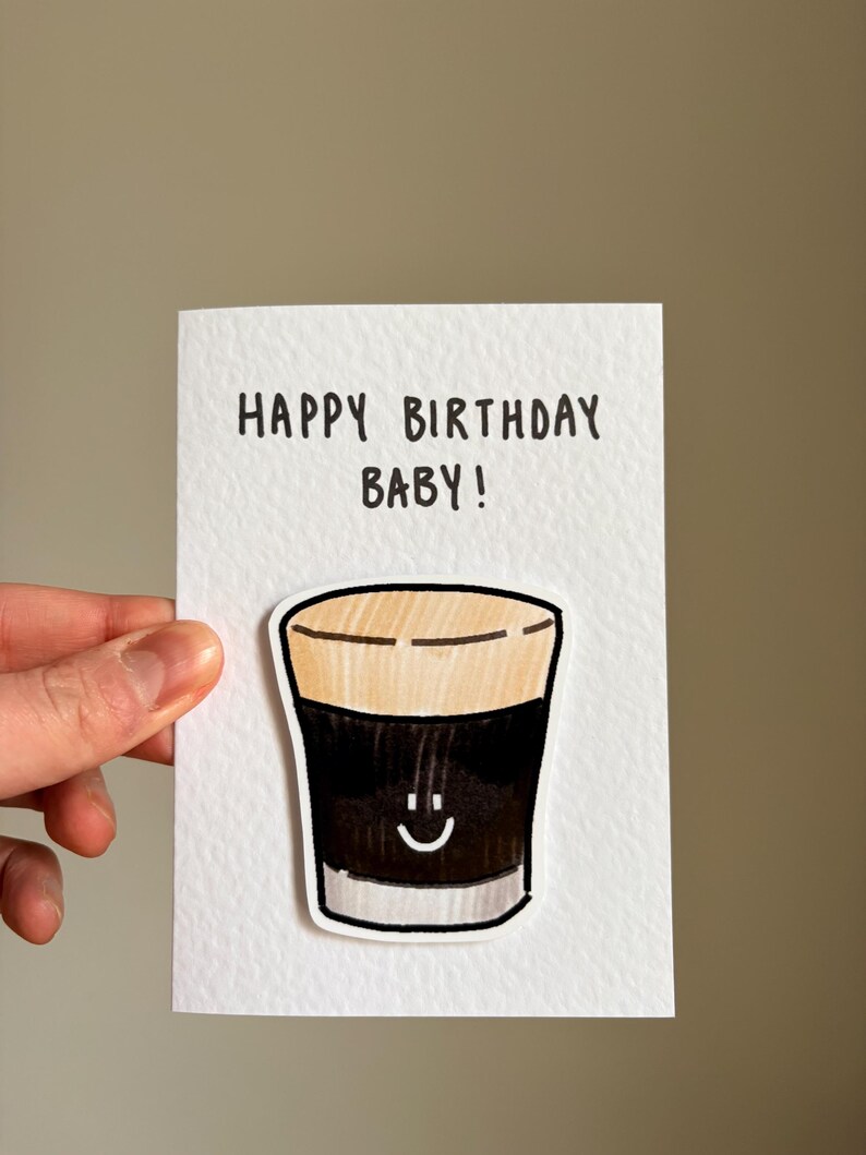 Puede incluir: Una tarjeta de felicitaci&oacute;n blanca con el texto "HAPPY BIRTHAY BABY!" en negro. La tarjeta presenta un dibujo de dibujos animados de una bebida oscura con una cara sonriente. La bebida tiene una parte superior de color canela y un cuerpo marr&oacute;n oscuro.