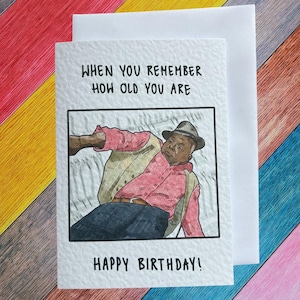 Op de afbeelding: Een verjaardagskaart met de tekst "WANNEER JE JE HERINNERT HOE OUD JE BENT" boven een illustratie van een man in een roze shirt en hoed. De woorden "HAPPY BIRTHDAY!" staan eronder.
