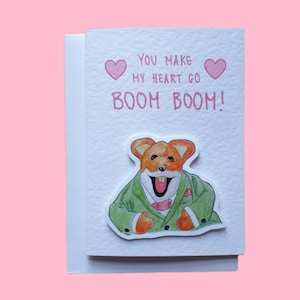 May include: A greeting card featuring a cartoon fox in a green suit and a pink bow tie. The card reads "You make my heart go BOOM BOOM!" with pink hearts on either side of the text.