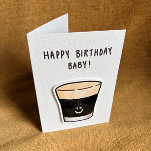 Puede incluir: Una tarjeta de felicitaci&oacute;n blanca con el texto "HAPPY BIRTHDAY BABY!" en negro. La tarjeta presenta una ilustraci&oacute;n de dibujos animados de una bebida oscura con una cara sonriente. La tarjeta est&aacute; sobre una superficie marr&oacute;n.