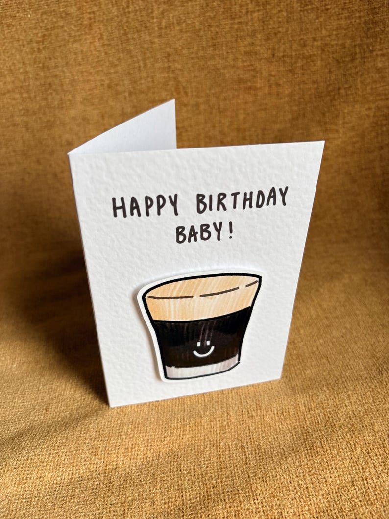 Puede incluir: Una tarjeta de felicitaci&oacute;n blanca con el texto "HAPPY BIRTHDAY BABY!" en negro. La tarjeta presenta una ilustraci&oacute;n de dibujos animados de una bebida oscura con una cara sonriente. La tarjeta est&aacute; sobre una superficie marr&oacute;n.