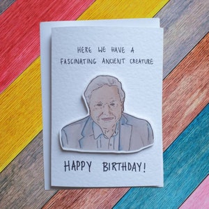 May include: A white card with a hand-drawn illustration of a man with grey hair and a light blue shirt. The text on the card reads "Here we have a fascinating ancient creature. Happy Birthday!"