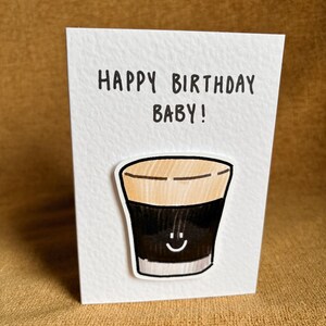 Puede incluir: Una tarjeta de felicitaci&oacute;n blanca con el texto "HAPPY BIRTHAY BABY!" en negro. La tarjeta presenta una ilustraci&oacute;n de dibujos animados de una bebida oscura con una cara sonriente. La bebida tiene una parte superior marr&oacute;n y un cuerpo negro.