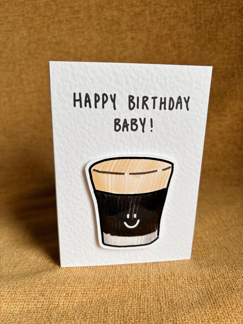 Puede incluir: Una tarjeta de felicitaci&oacute;n blanca con el texto "HAPPY BIRTHAY BABY!" en negro. La tarjeta presenta una ilustraci&oacute;n de dibujos animados de una bebida oscura con una cara sonriente. La bebida tiene una parte superior marr&oacute;n y un cuerpo negro.