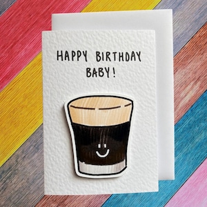 Puede incluir: Una tarjeta de felicitaci&oacute;n blanca con el texto "HAPPY BIRTHDAY BABY!" en negro. Un dibujo animado de una cerveza negra oscura con una cara sonriente est&aacute; en la tarjeta. La tarjeta est&aacute; sobre una superficie de madera multicolor.
