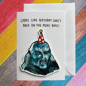 May include: A white greeting card with the text "LOOKS LIKE BIRTHDAY CAKE'S BACK ON THE MENU BOYS!" features a blue-toned illustration of a creature wearing a party hat. The card is paired with a white envelope.