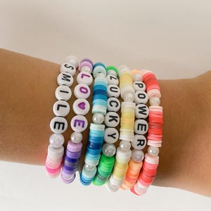 Op de afbeelding: Een verzameling kleurrijke kralenarmbanden met woorden als "SMILE", "LOVE", "LUCKY" en "POWER". De armbanden hebben een verscheidenheid aan kleuren, waaronder roze, paars, blauw, groen, geel, oranje en rood, met witte letterkralen en parelaccenten.