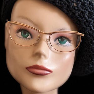 Peut inclure: Tête de mannequin portant des lunettes à monture dorée et un béret noir en crochet sur fond noir. Les lunettes ont une forme œil de chat. Le mannequin a les yeux verts et du rouge à lèvres rouge.