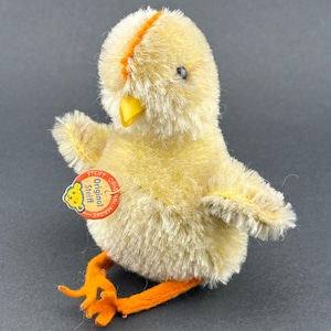 Könnte beinhalten: Ein Vintage-Plüschküken-Spielzeug mit gelbem Fell, orangefarbenen Füßen und einem gelben Schnabel. Das Küken hat ein rot-weißes Etikett mit der Aufschrift "Steiff Original". Das Spielzeug befindet sich vor einem dunkelgrauen Hintergrund.