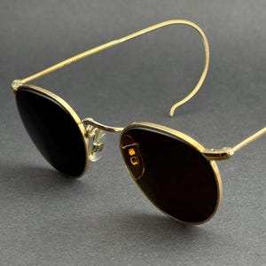 Peut inclure: Lunettes de soleil à monture dorée avec verres foncés. Les lunettes ont une forme ovale classique et un design minimaliste, avec des branches fines et incurvées. Les verres semblent teintés, offrant de l'ombre. L'esthétique générale est élégante et vintage.