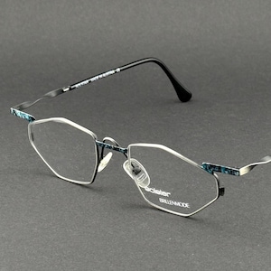 Könnte beinhalten: Brille mit einem silberfarbenen, kantigen, rahmenlosen Gestell mit blaugrün-schwarzen Mustern an den Scharnieren. Die Bügel haben ein markantes, gewelltes Metalldesign und das rechte Glas ist mit dem Text "solster BRILLENMODE" graviert.