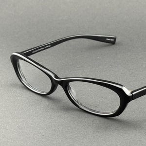 Könnte beinhalten: Schwarze Brille mit weißem Rand. Die Katzenaugen-Rahmen haben klare Gläser. Die Bügel sind schwarz mit einem weißen Streifen. Der Text "OLIVER PEOPLES" ist auf dem linken Bügel aufgedruckt. Ein modisches Accessoire.