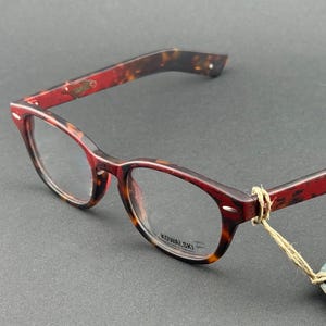 Könnte beinhalten: Brille mit Schildpattmuster auf Rahmen und Bügeln. Die Rahmen sind eine Mischung aus roten und braunen Farbtönen. Die Gläser sind klar. Ein kleines Etikett ist am Rahmen befestigt. Der Markenname "KOWALSKI" ist sichtbar.