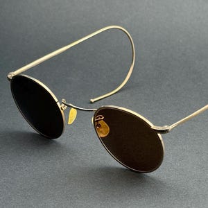Peut inclure: Lunettes de soleil en métal doré avec des verres ovales. Les montures ont un design minimaliste, avec un fil fin s'étendant des verres pour s'enrouler autour des oreilles. Les verres sont d'une couleur sombre et opaque, offrant un look élégant et moderne.