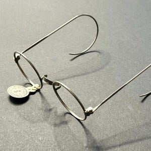 Puede incluir: Gafas plateadas con montura de alambre, lentes redondas y patillas curvas. Una pequeña etiqueta circular está adherida a la montura. Las gafas se presentan sobre un fondo oscuro, resaltando su delicado diseño.
