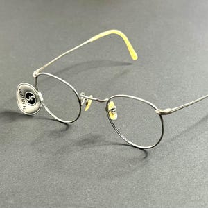 Peut inclure: Lunettes à monture argentée avec verres ovales et embouts de branches jaunes. Les lunettes ont une petite étiquette ronde attachée à la monture avec le texte "PLATININ" et un logo. Elles sont posées sur un fond gris foncé.