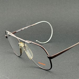 Könnte beinhalten: Braune Metall-Aviator-Brille mit silbernen, gebogenen Bügeln. Die Brille hat einen Doppelsteg und klare Gläser. Der Markenname "MENRAD" ist auf dem Rahmen sichtbar. Vor dunklem Hintergrund.