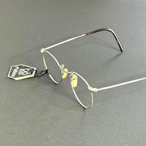 Peut inclure: Lunettes à monture argentée avec verres ovales et branches à motif écaille de tortue marron. Une petite étiquette est attachée à la monture. Les lunettes sont posées sur un fond gris.