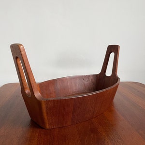 Può includere: Una ciotola di legno a forma di barca con due manici alti e curvi. La ciotola è realizzata in un ricco legno marrone-rossastro e poggia su una superficie di legno abbinata. Le maniglie hanno un ritaglio rettangolare. Questa ciotola decorativa è ideale per servire o esporre.