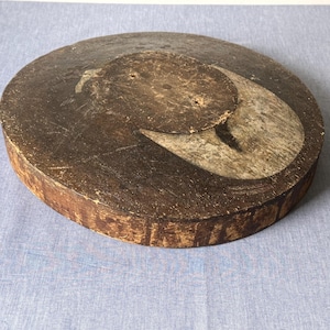 Pode incluir: Uma tábua de cortar redonda de madeira com uma superfície castanha escura e um design curvo mais claro. A tábua tem uma peça circular presa no topo. As bordas mostram o grão natural da madeira. Este item de cozinha rústico é provavelmente usado para preparar alimentos.